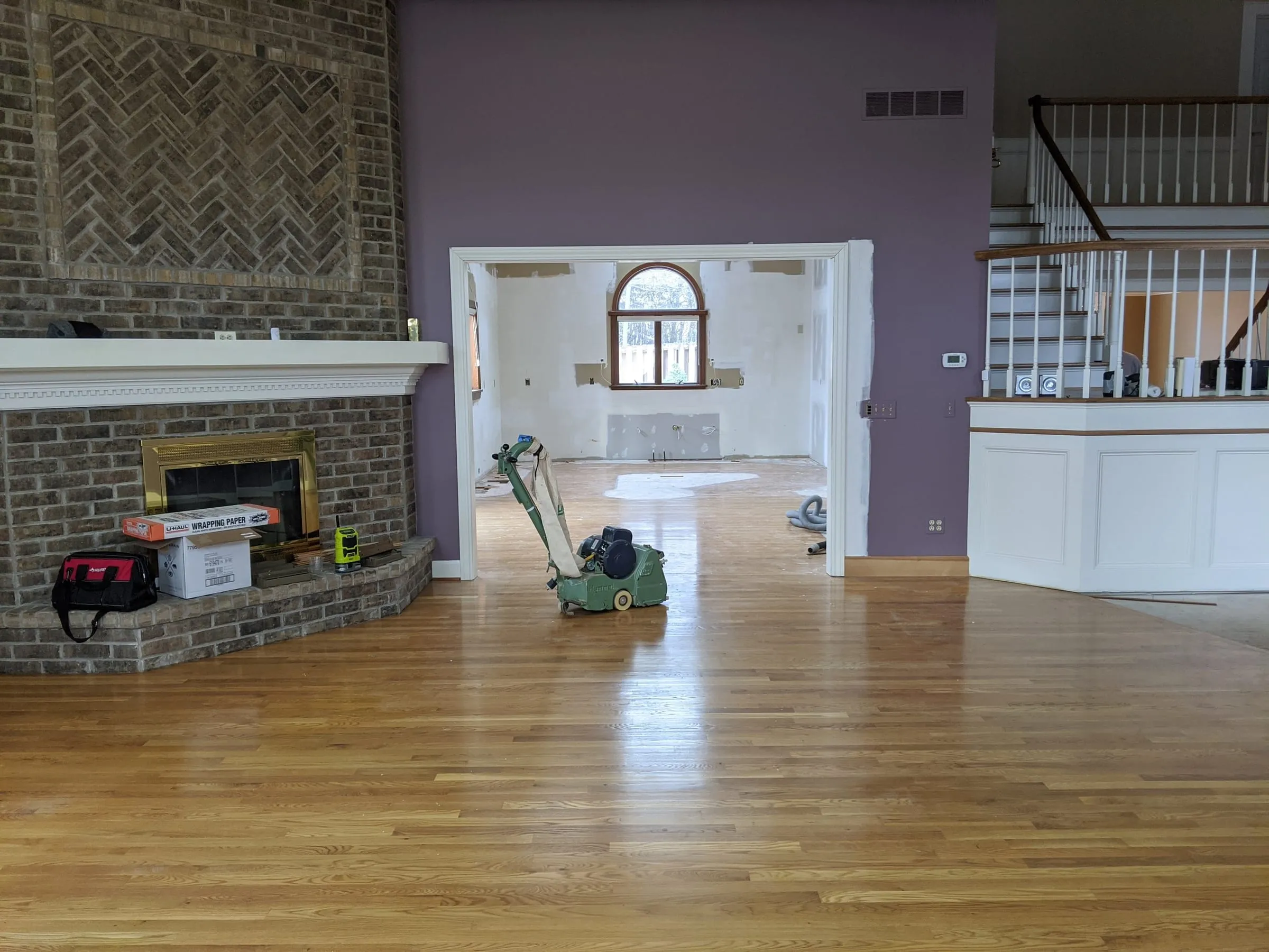 Open living room with brick fireplace and oak floors midway through refinishing